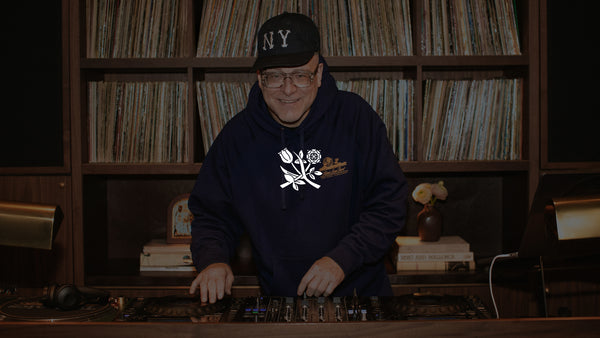 Person wearing a black hoodie with a white logo and a cap, standing in front of a record player and vinyl records.