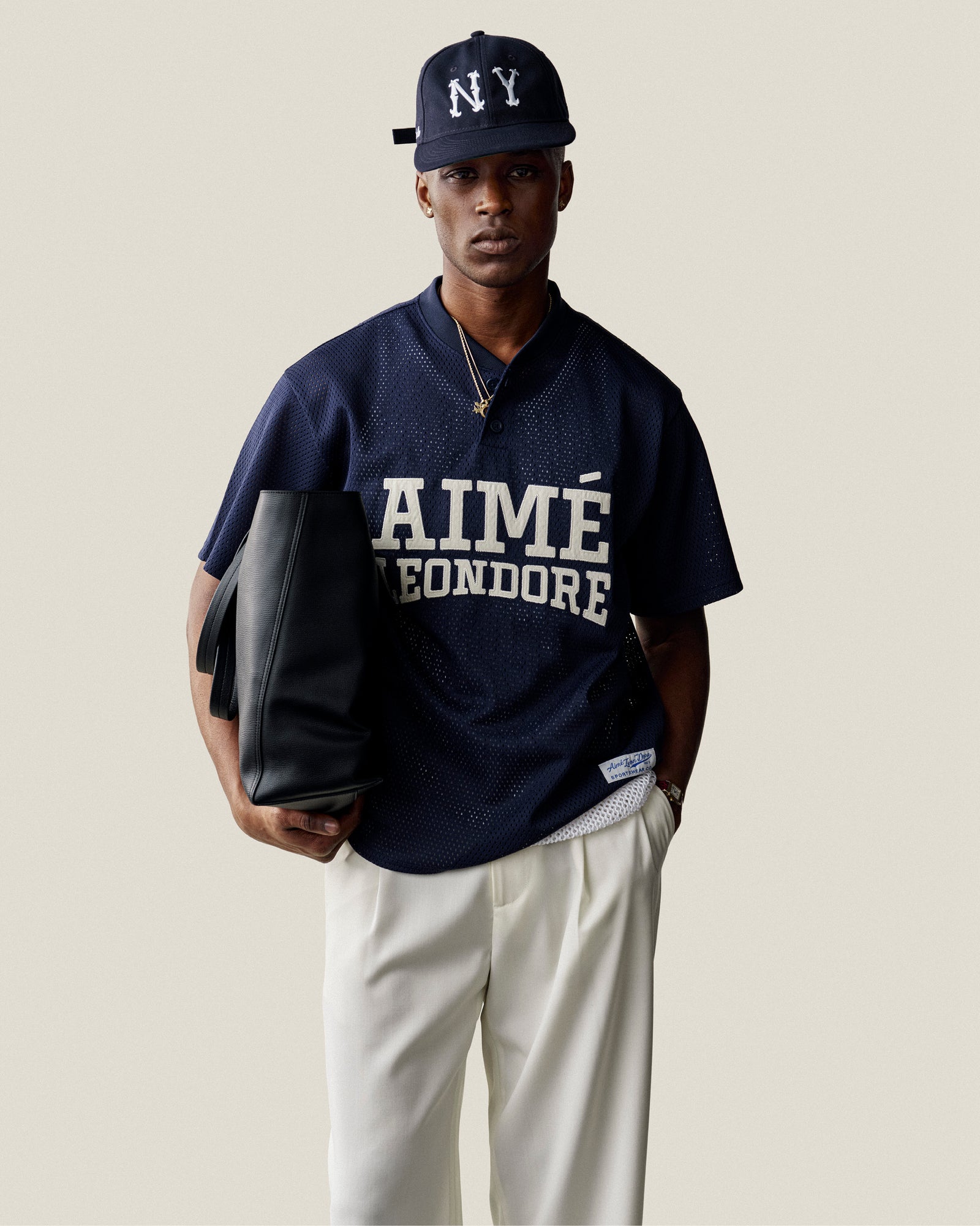 Man wears ALD / New Era Highlanders Retro Fit Hat, navy Aimé Leon Dore jersey, white pants, and holds a black bag.