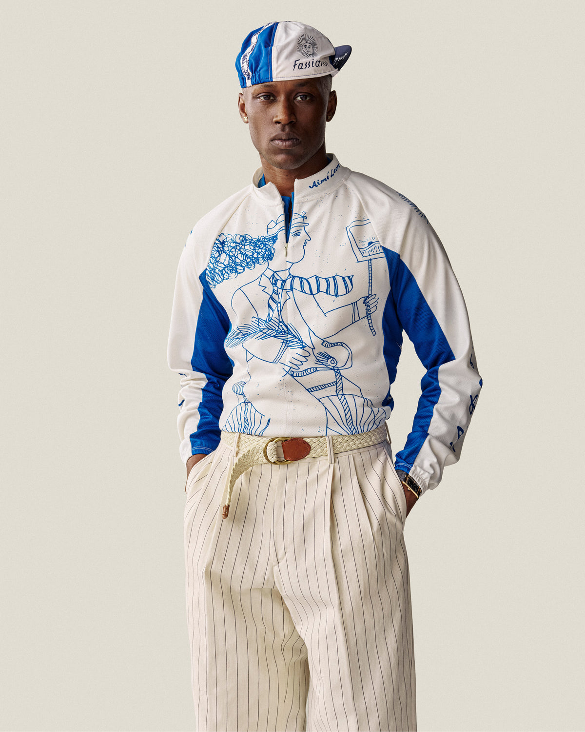 Man wearing a Fassianos Cycling Cap, graphic zip-up top, and beige pinstripe pants with hands in pockets.