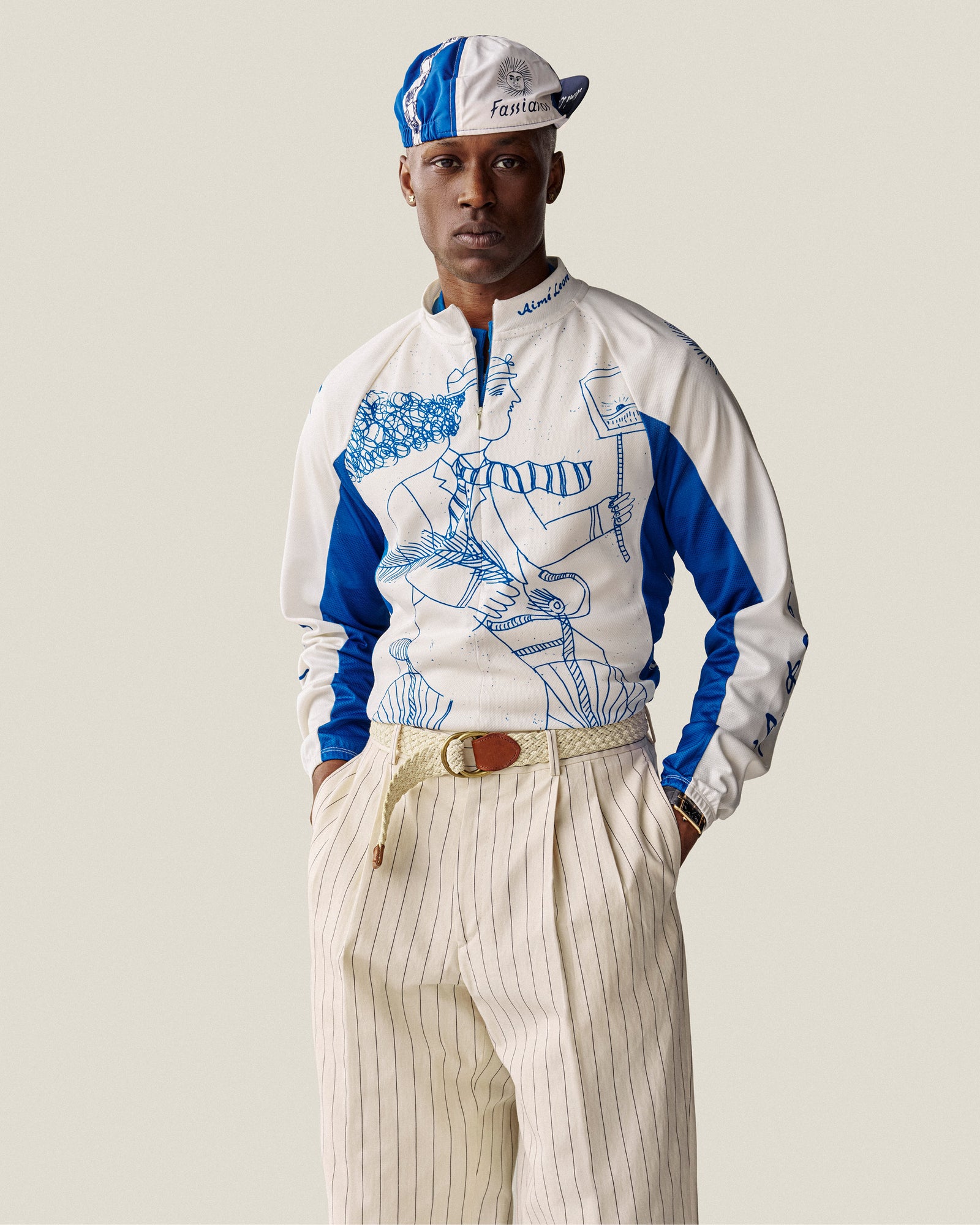Man wearing a Fassianos Cycling Cap, graphic zip-up top, and beige pinstripe pants with hands in pockets.