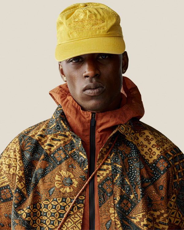 A man in a Sun Faded Unisphere Hat and an earthy patterned jacket, looking directly at the camera.