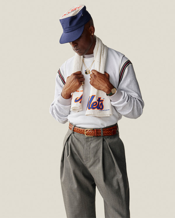 Person wearing a Mets shirt, gray pants, scarf, necklaces, watch, braided belt, and an Aimé Painters Hat, looking down.