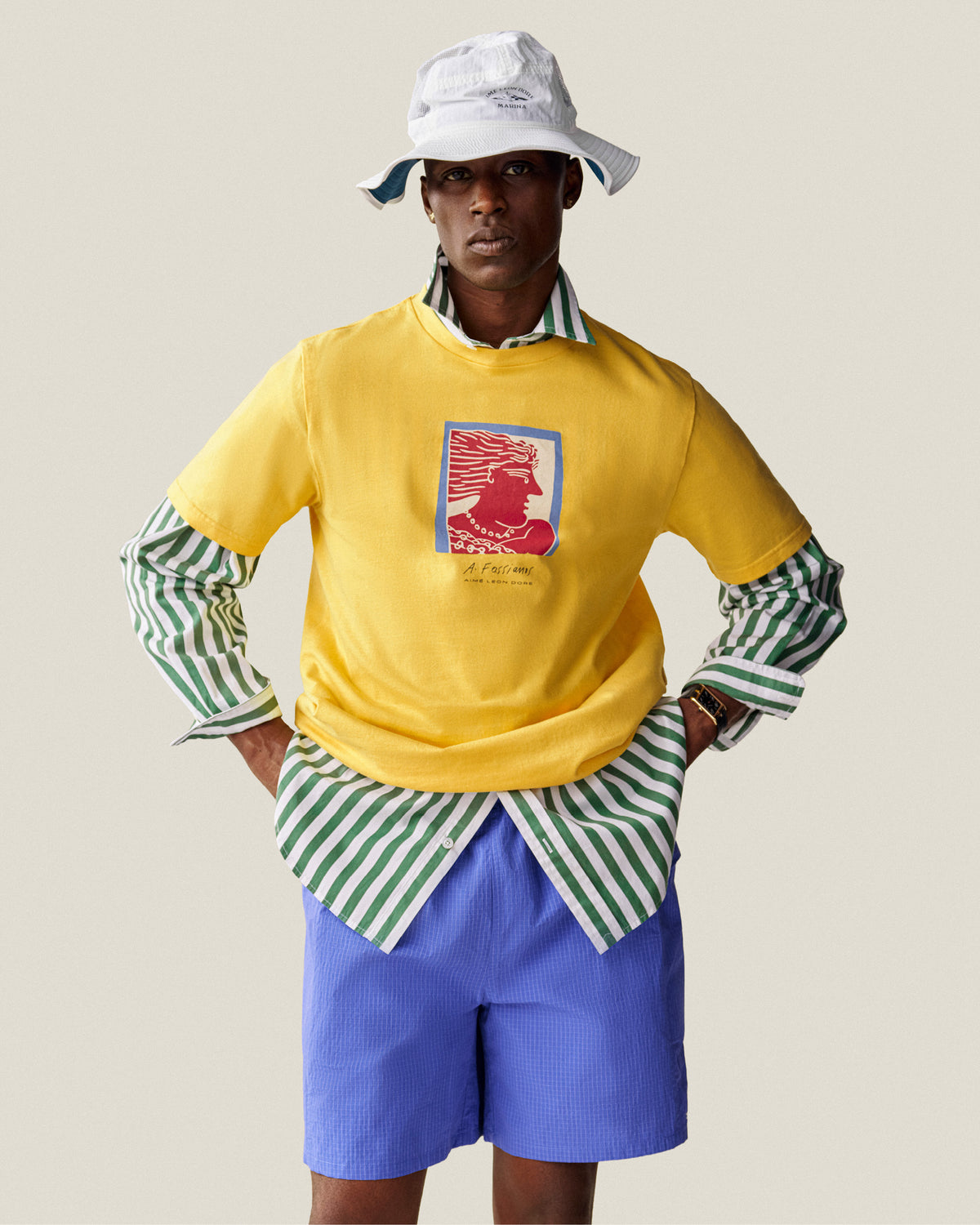 A man wears a white bucket hat, yellow graphic tee, Striped Leon Overshirt, and blue shorts against a plain background.