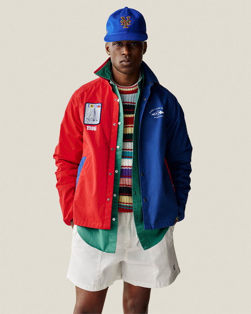 Man wearing a blue and red jacket, striped crewneck sweater, green shirt, white shorts, and a blue baseball cap.