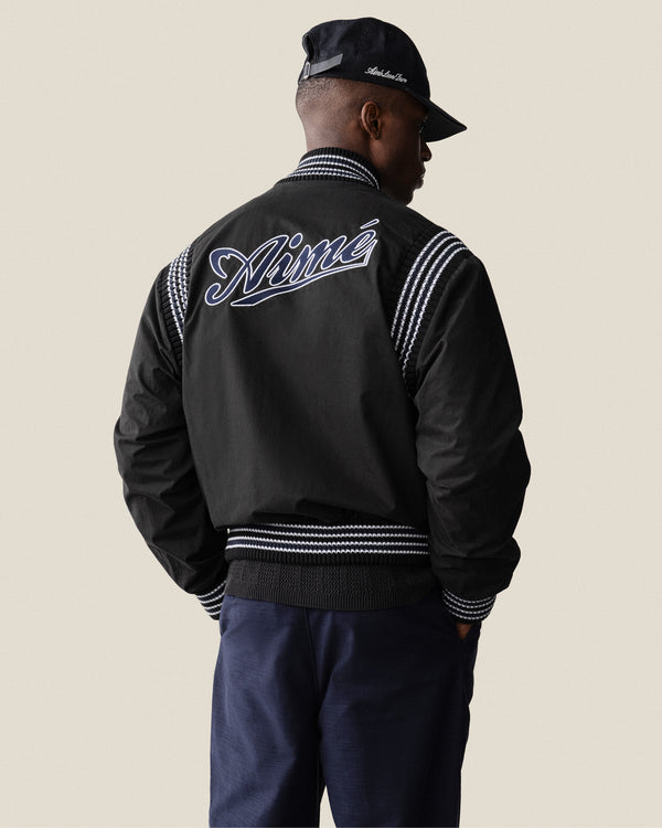 Man wears Reversible Knit Bomber Jacket with Nimé on the back, black cap, dark pants, facing away.
