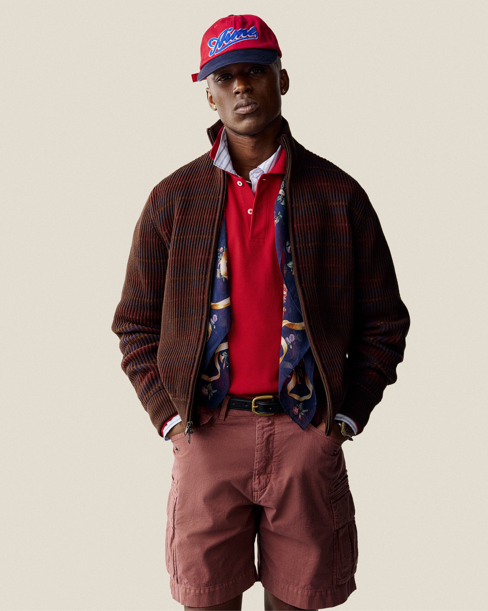 A man in a red cap and Multi Color Knit Track Sweater stands against a plain background.