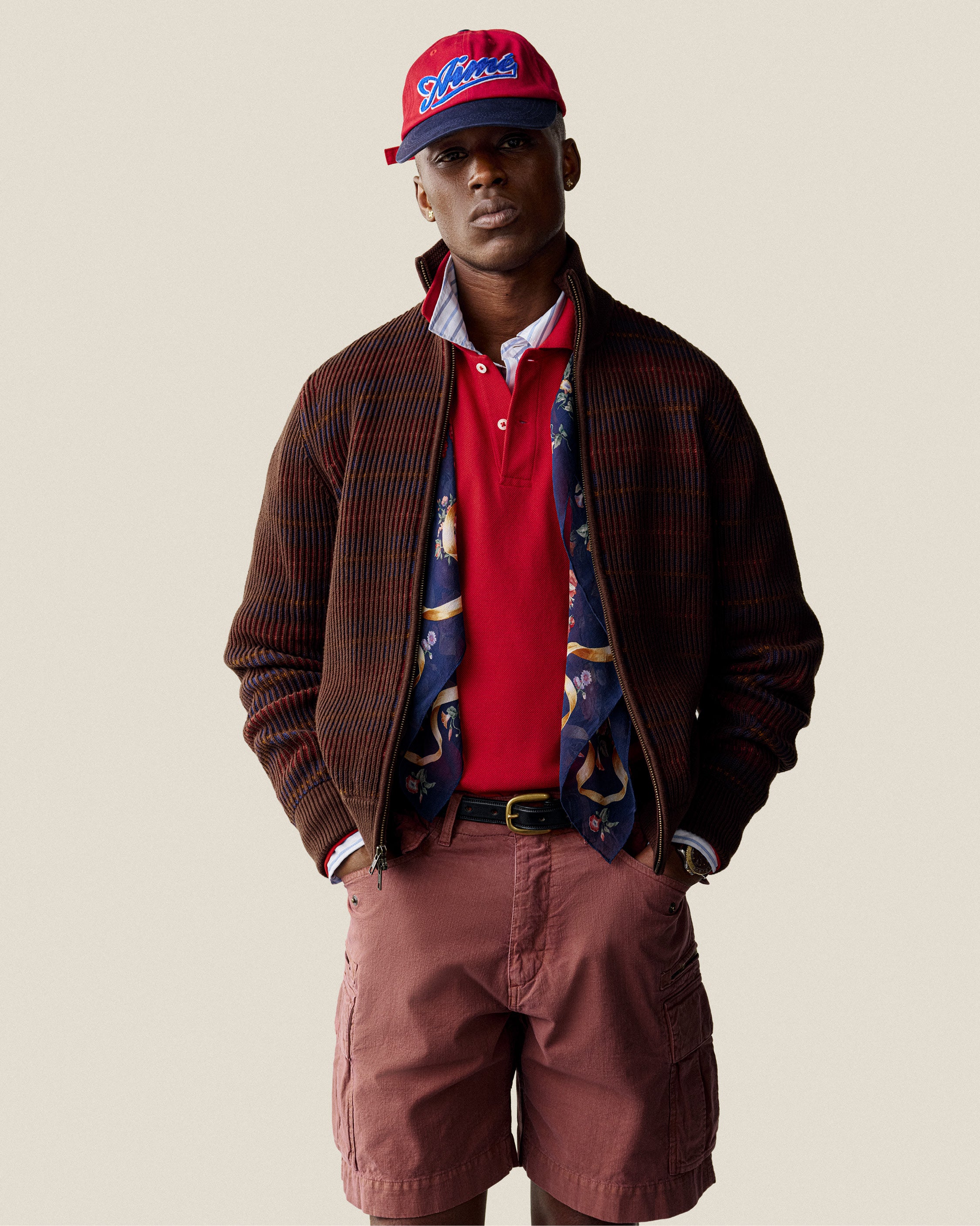 A man in a red cap and Multi Color Knit Track Sweater stands against a plain background.