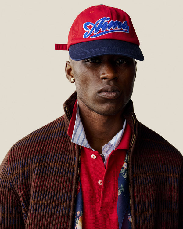 Man wearing a Cycling Logo Hat, red shirt, striped jacket, and gold earring, looking serious against a plain background.