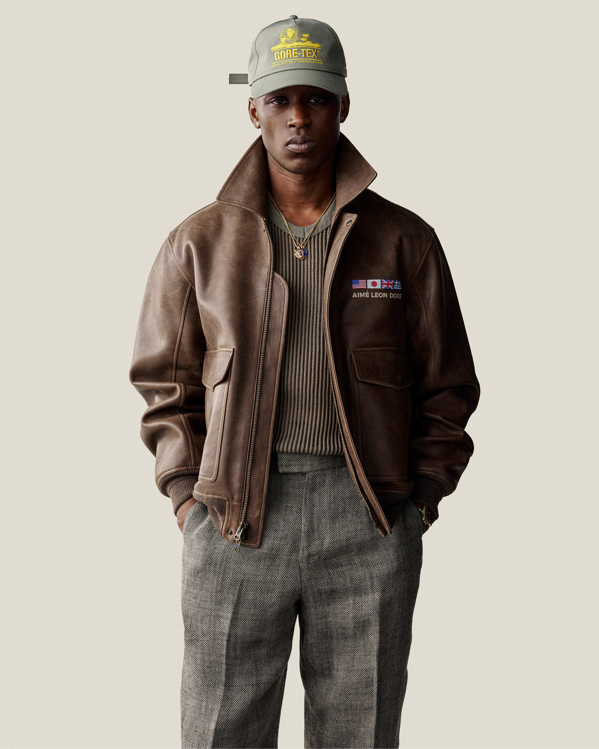 A man in the World's Borough Leather Flight Jacket, gray pants, and a green GORE-TEX cap stands by a plain background.