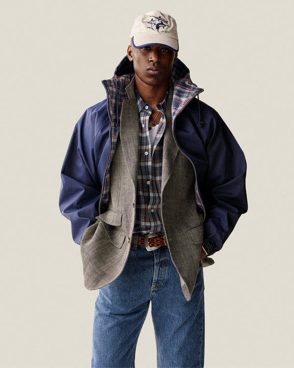 A man wears a Washed Madras Shirt, gray blazer, blue jacket, jeans, and a white baseball cap with hands in pockets.
