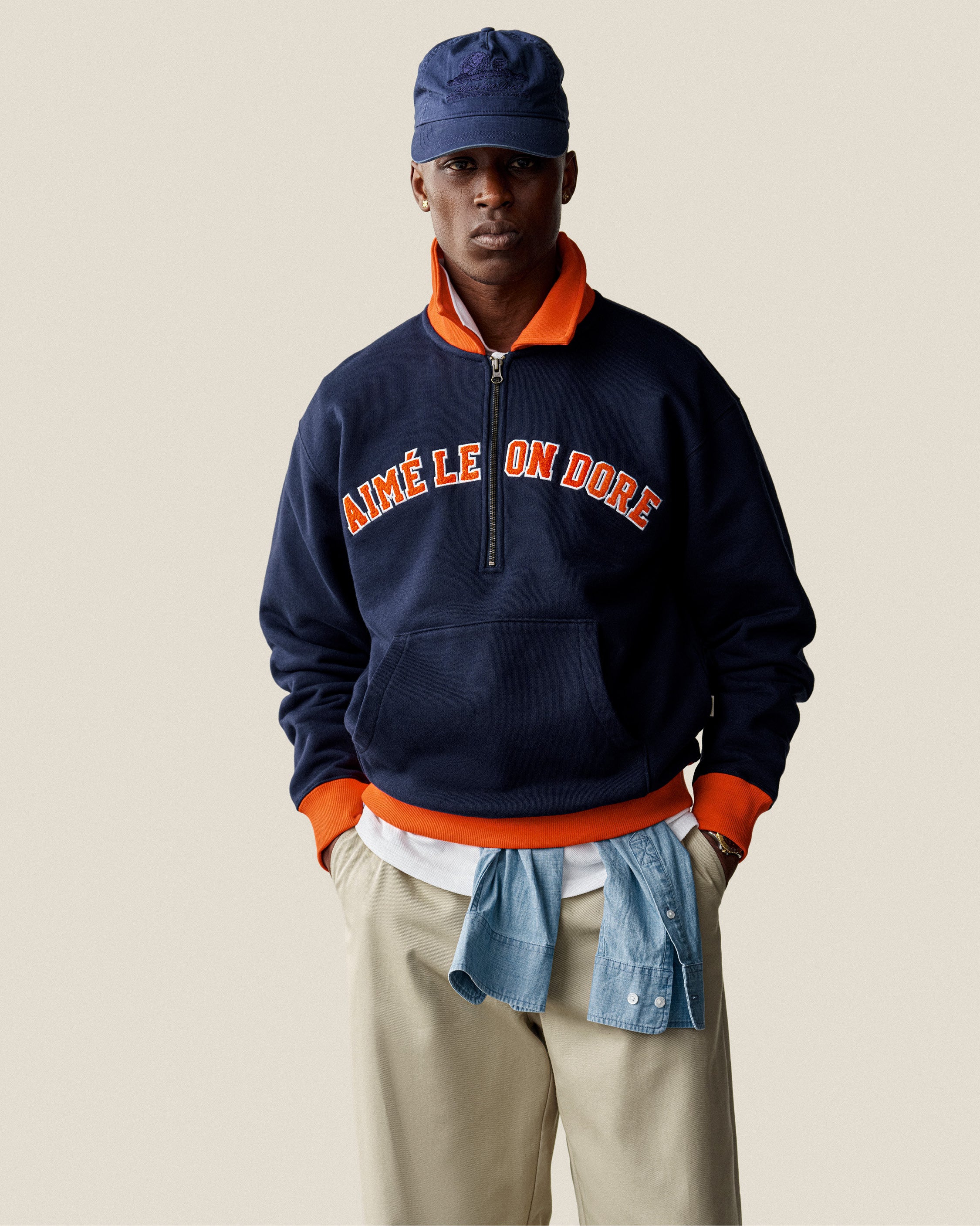 A man in a navy Collegiate Quarter Zip Pullover with AIMÉ LE ON DORE text, khaki pants, and a denim shirt at the waist.