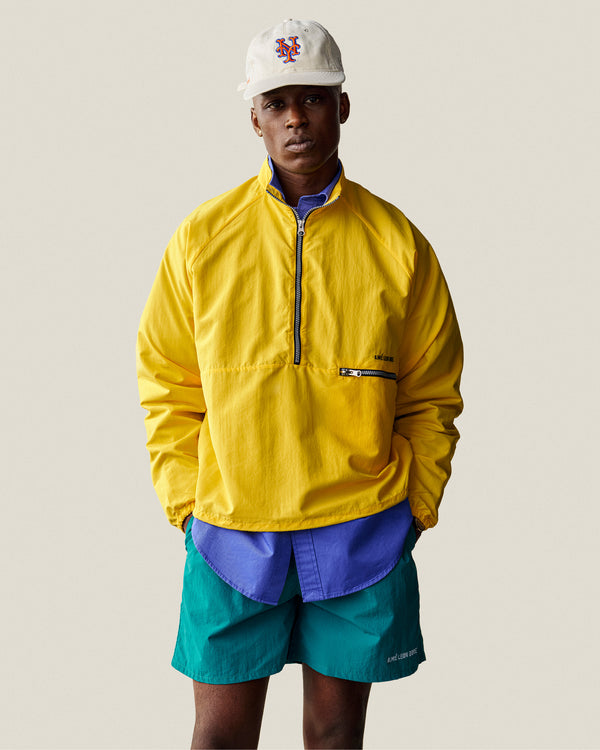 Man wearing a Leon Overshirt, yellow pullover, teal shorts, and white cap against a plain background.