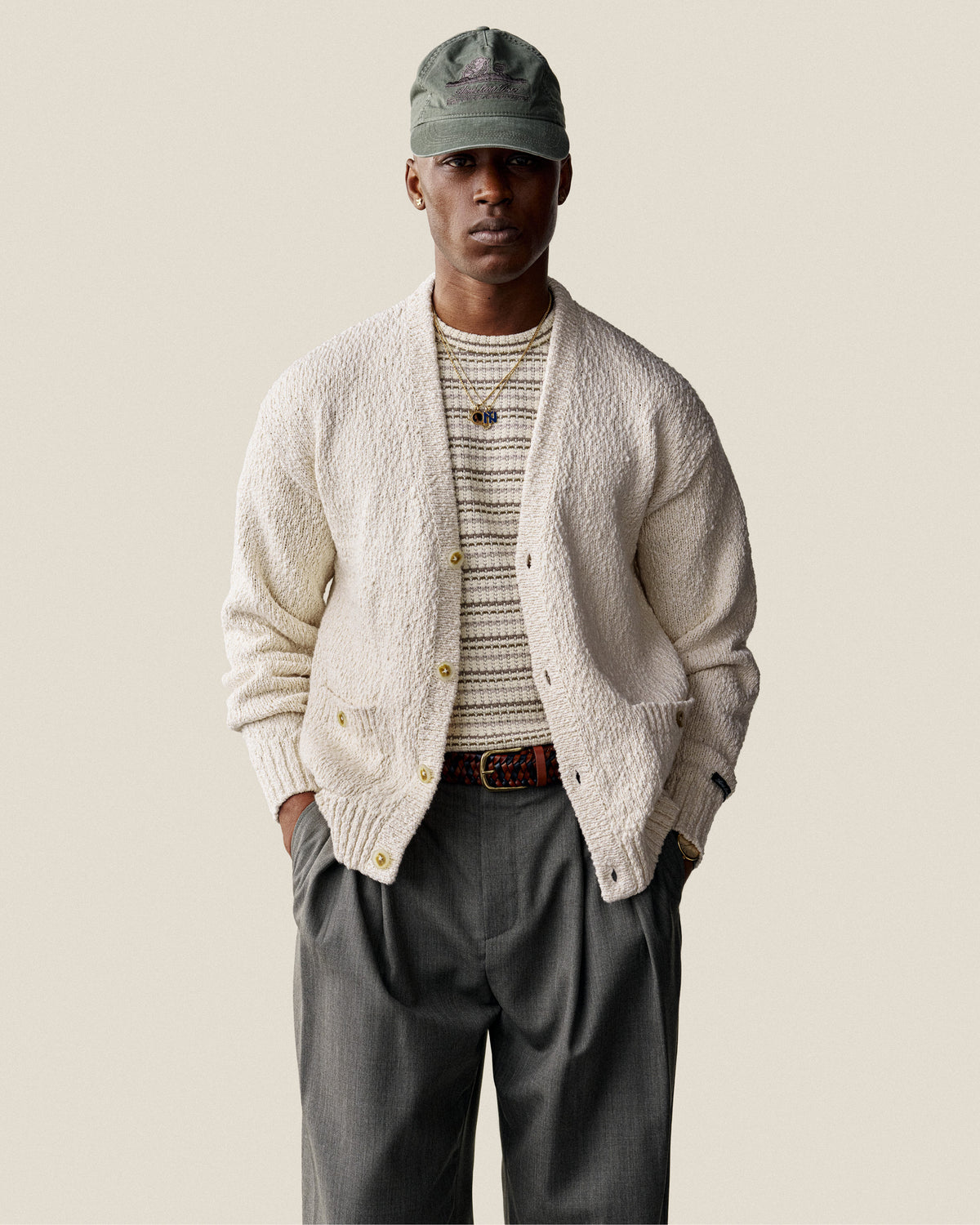 Man wearing a Sun Faded Unisphere Hat, cream cardigan, striped shirt, and gray pants stands with hands in pockets.