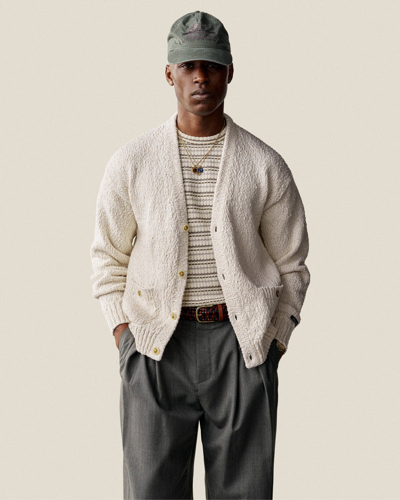 Man wearing a Sun Faded Unisphere Hat, cream cardigan, striped shirt, and gray pants stands with hands in pockets.