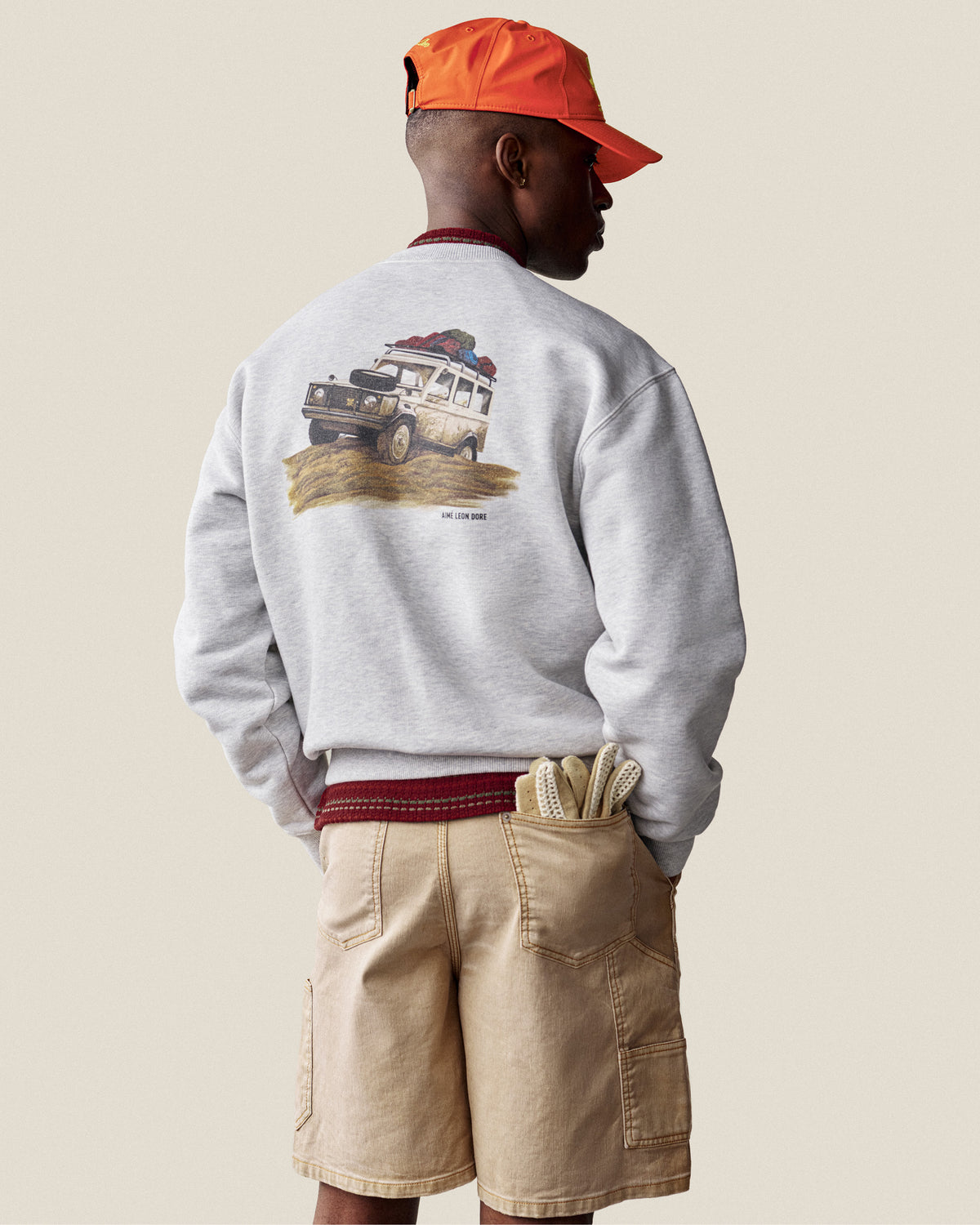 Man in a Safari Crewneck Sweatshirt, tan shorts, orange cap, and gardening gloves in pocket, facing away.