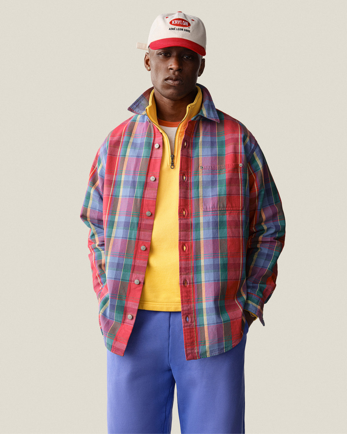 A person in Sun Faded Sweatpants, red plaid shirt, yellow top, and white cap stands against a plain background.