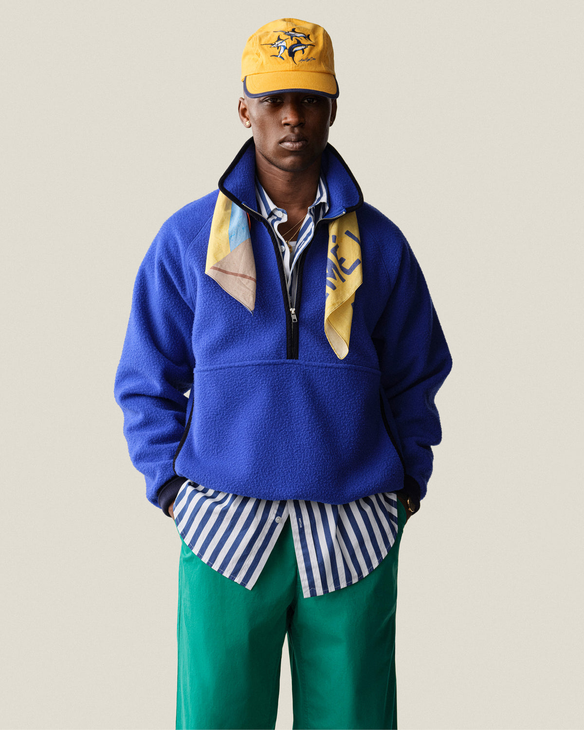 A person in a Striped Leon Overshirt, blue fleece pullover, green pants, yellow scarf, and yellow cap stands facing forward.