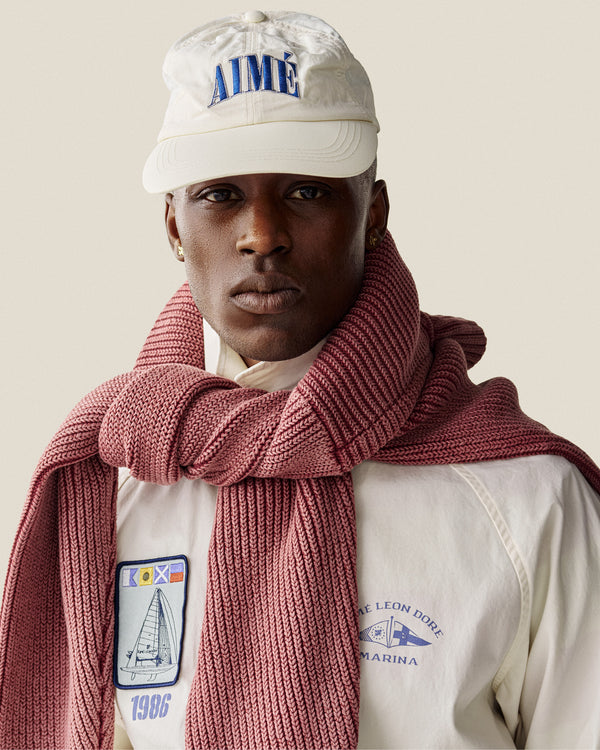 A man in a white Nylon Crusher Hat, white jacket with patches, and a pink knit scarf draped over his shoulders.