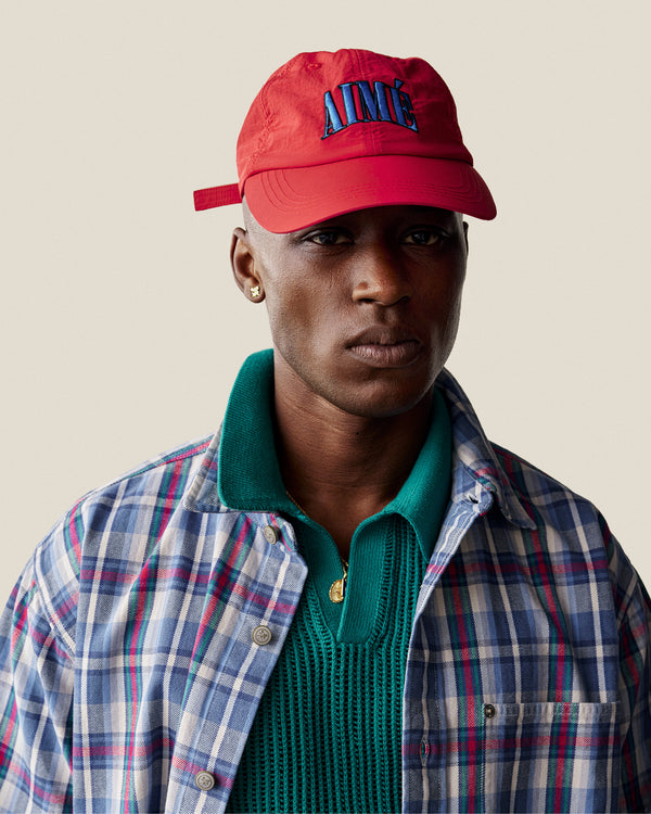 A person wearing a Nylon Crusher Hat, plaid shirt, green knit sweater, and gold earrings looks at the camera neutrally.