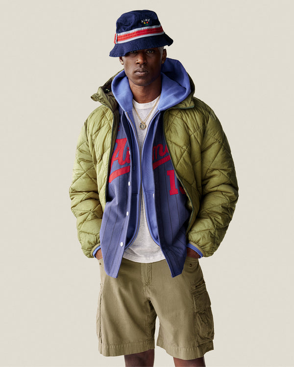 A man wearing a bucket hat, blue hoodie, jersey, khaki cargo shorts, and a Nylon Quilted Unisphere Jacket stands against a plain background.