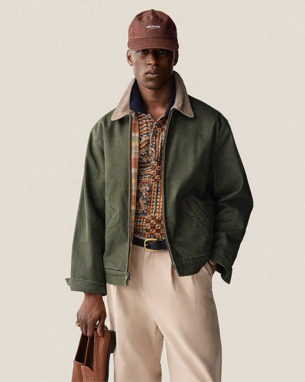 A man in a green jacket, patterned shirt, beige pants, and a Vintage Micro Logo Hat holds a brown bag.