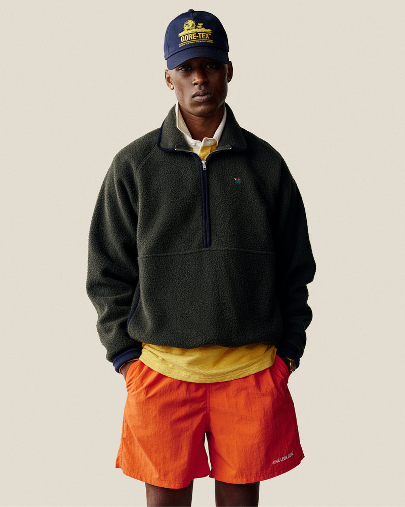Man in GORE-TEX Unisphere Hat, dark fleece pullover, yellow shirt, and orange shorts stands against plain background.