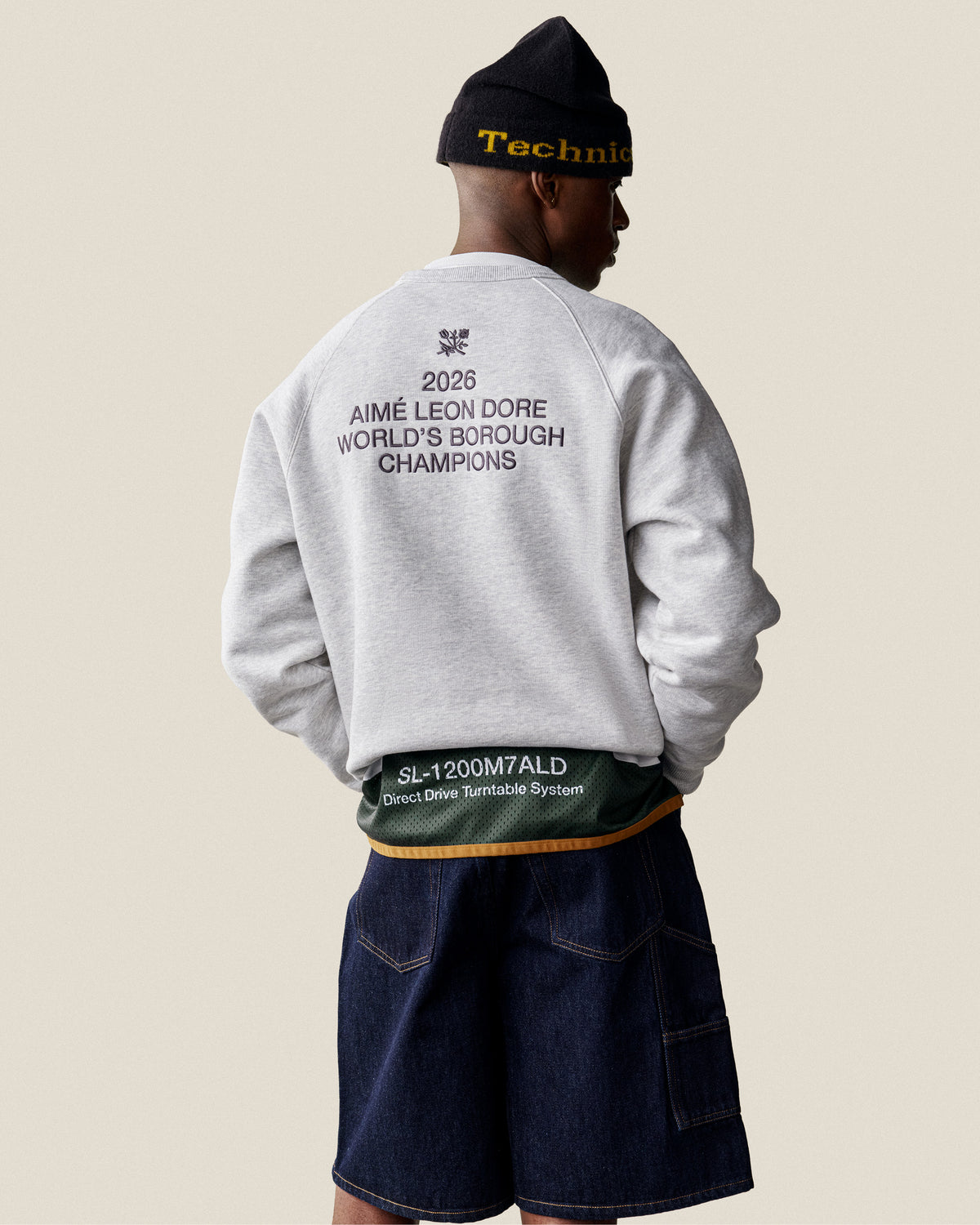 Man wearing a beanie, denim shorts, and an ALD / Technics World Champions Crewneck gray sweatshirt.