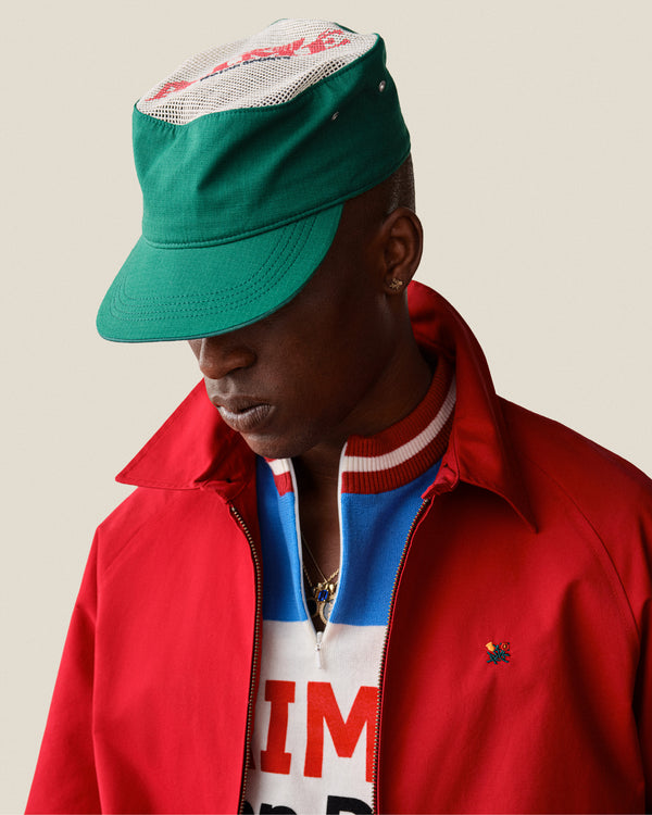Wearing the Aimé Painters Hat, a person in a red jacket and blue-and-white shirt looks down against a plain background.