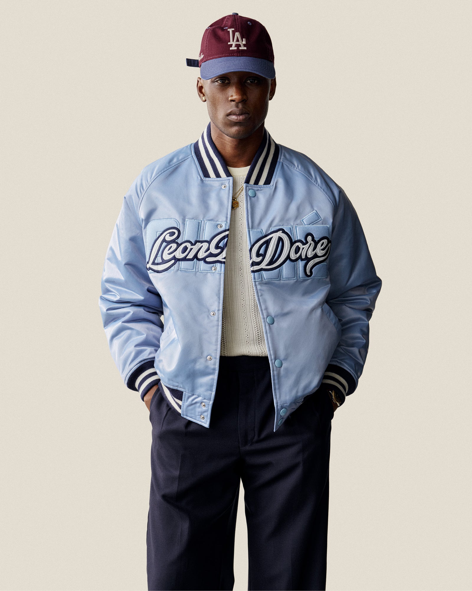 Man in an Overlay Logo Satin Varsity Jacket, white sweater, dark pants, and burgundy LA cap against a plain background.
