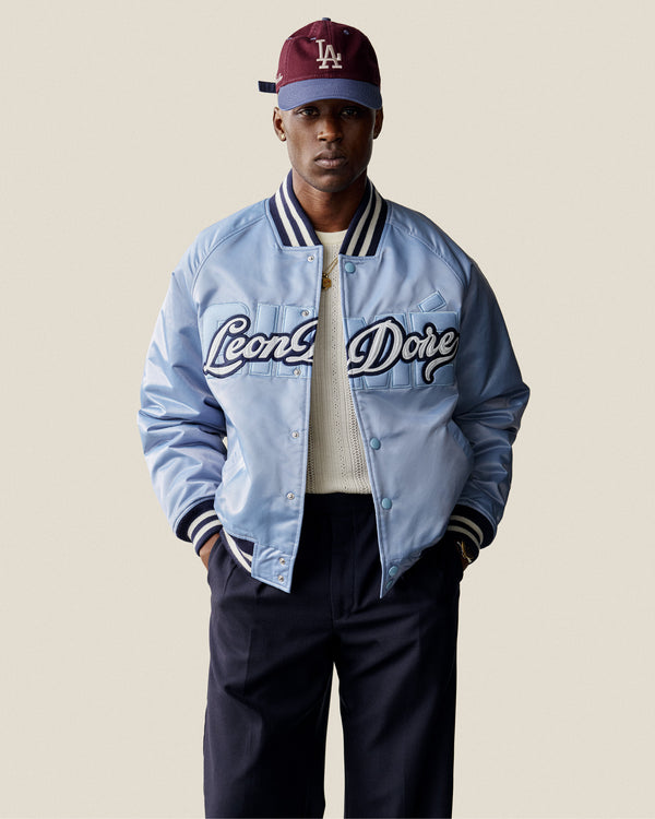 A man in a light blue Leon Dore varsity jacket, Merino Wool Eyelet Tee, dark pants, and burgundy LA cap.
