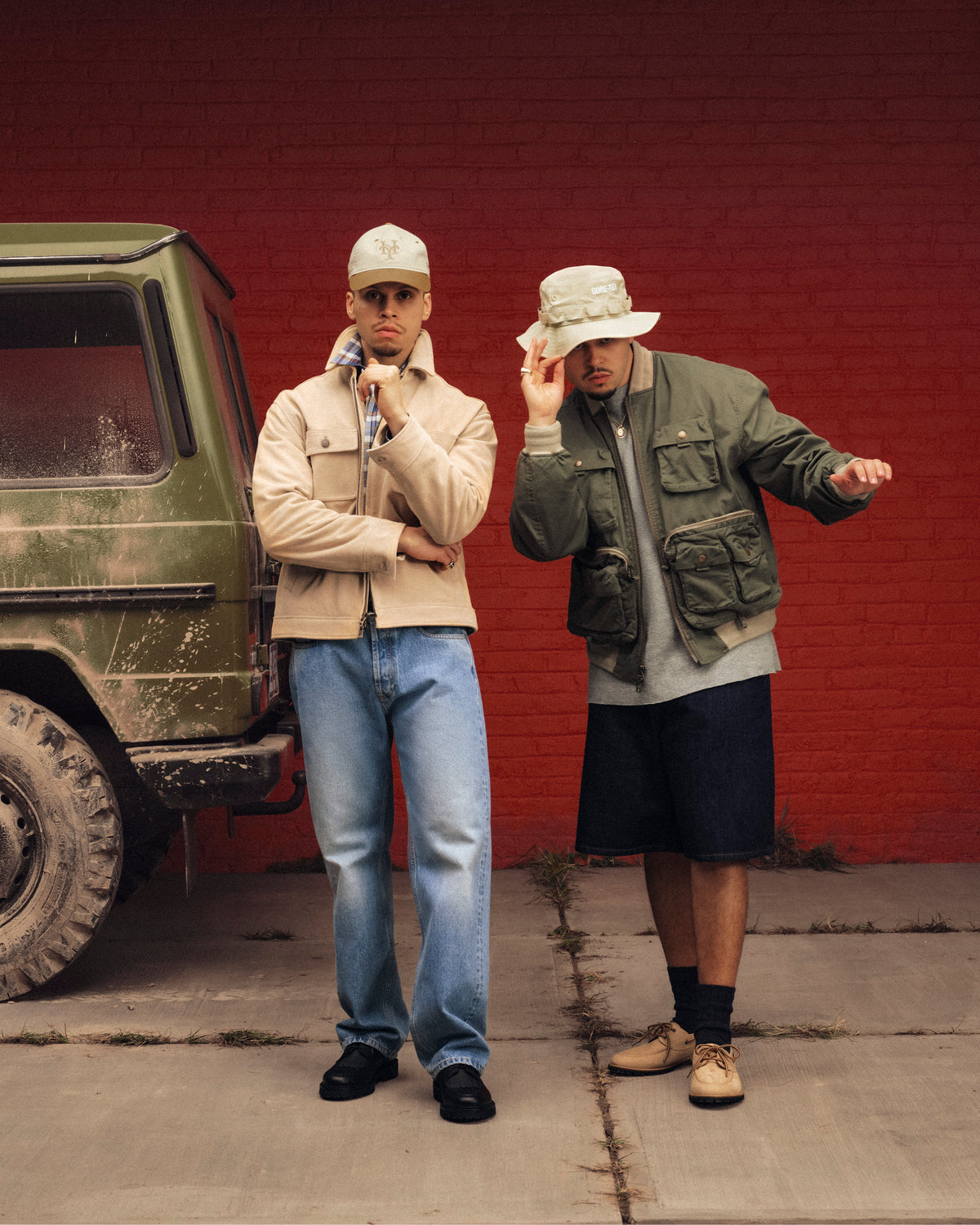 Two men in Safari Utility Bomber jackets and hats pose by a muddy green SUV against a red brick wall.