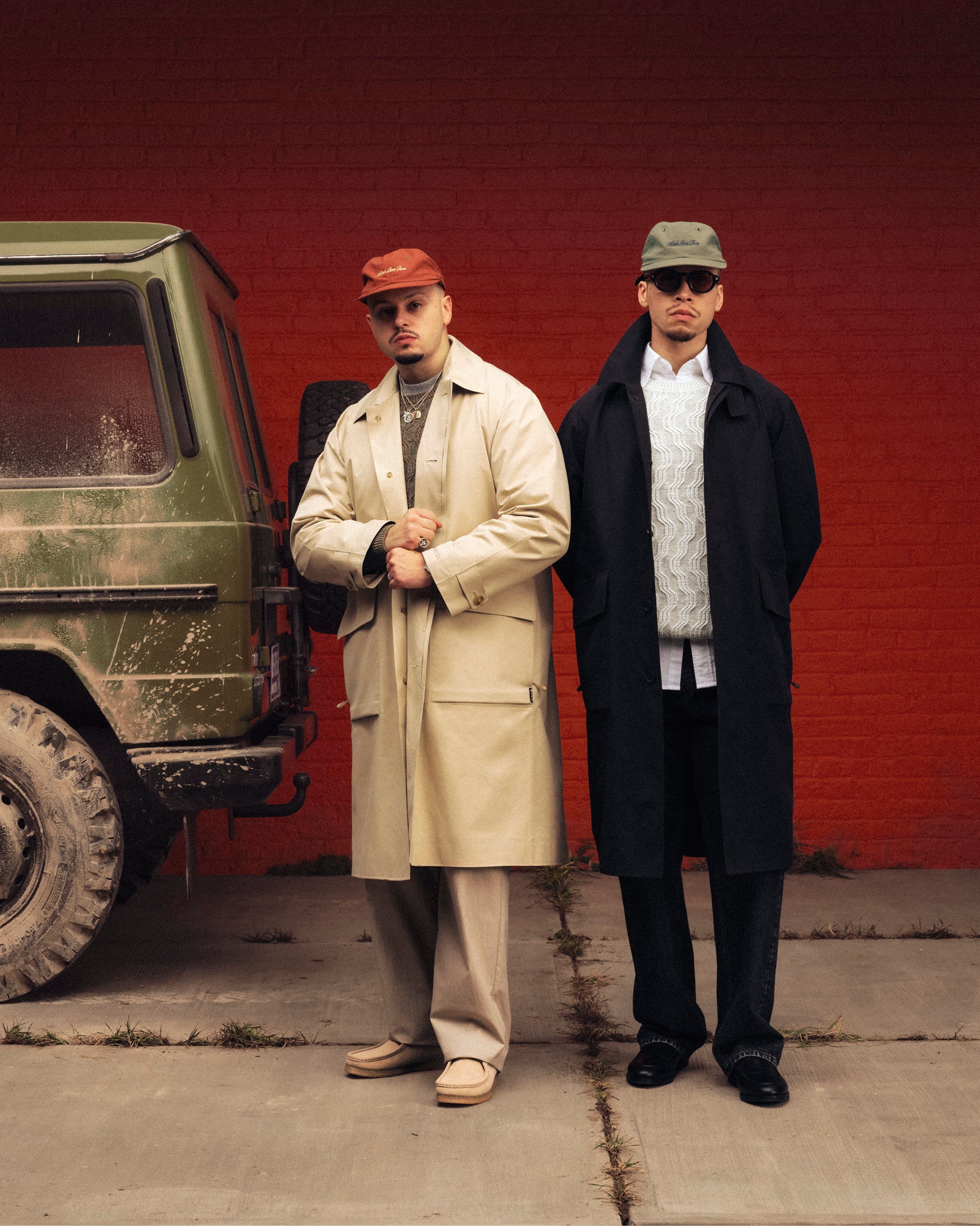 Two men in GORE-TEX Balmacaan Coats and hats pose by a green SUV against a red brick wall for a fashion photo.
