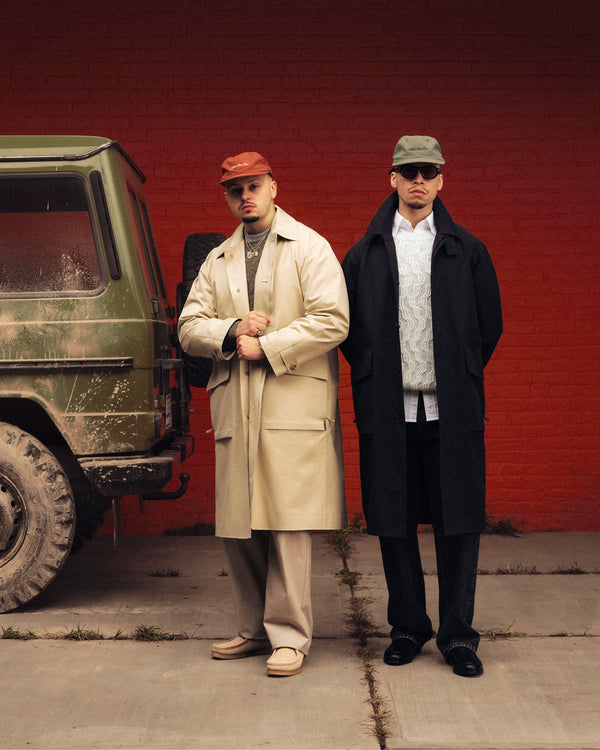 Two men in stylish coats and Script Sport Hats stand by a muddy green SUV, in front of a red brick wall.