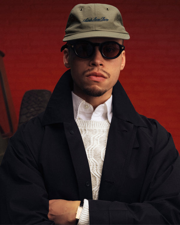Man in a Script Sport Hat, black coat, white sweater, and sunglasses stands against a red background looking serious.