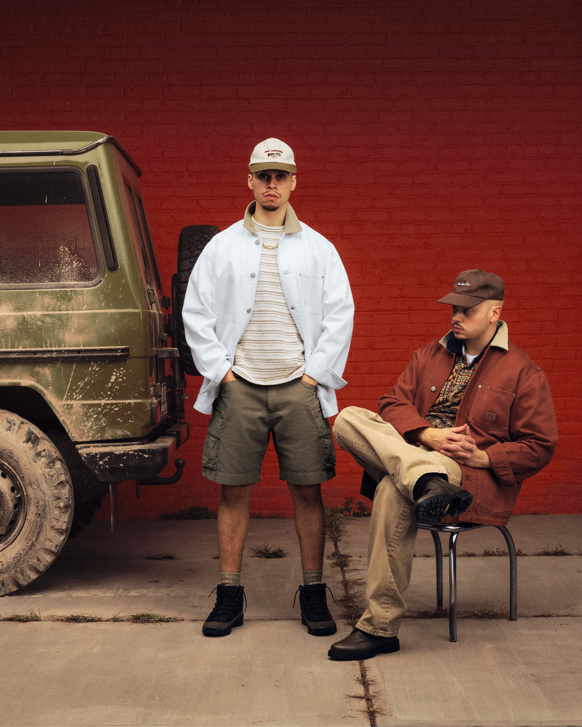 Two men in Denim Barn Jackets pose by a muddy green SUV and red brick wall; one standing, one sitting on a chair.