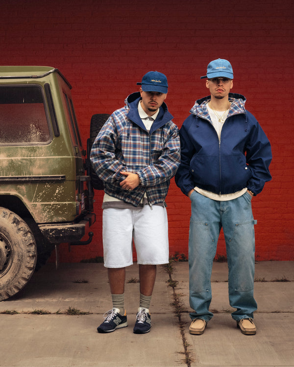 Two men, one in Light Wash Denim Shorts, stand beside a muddy green SUV and a red brick wall.