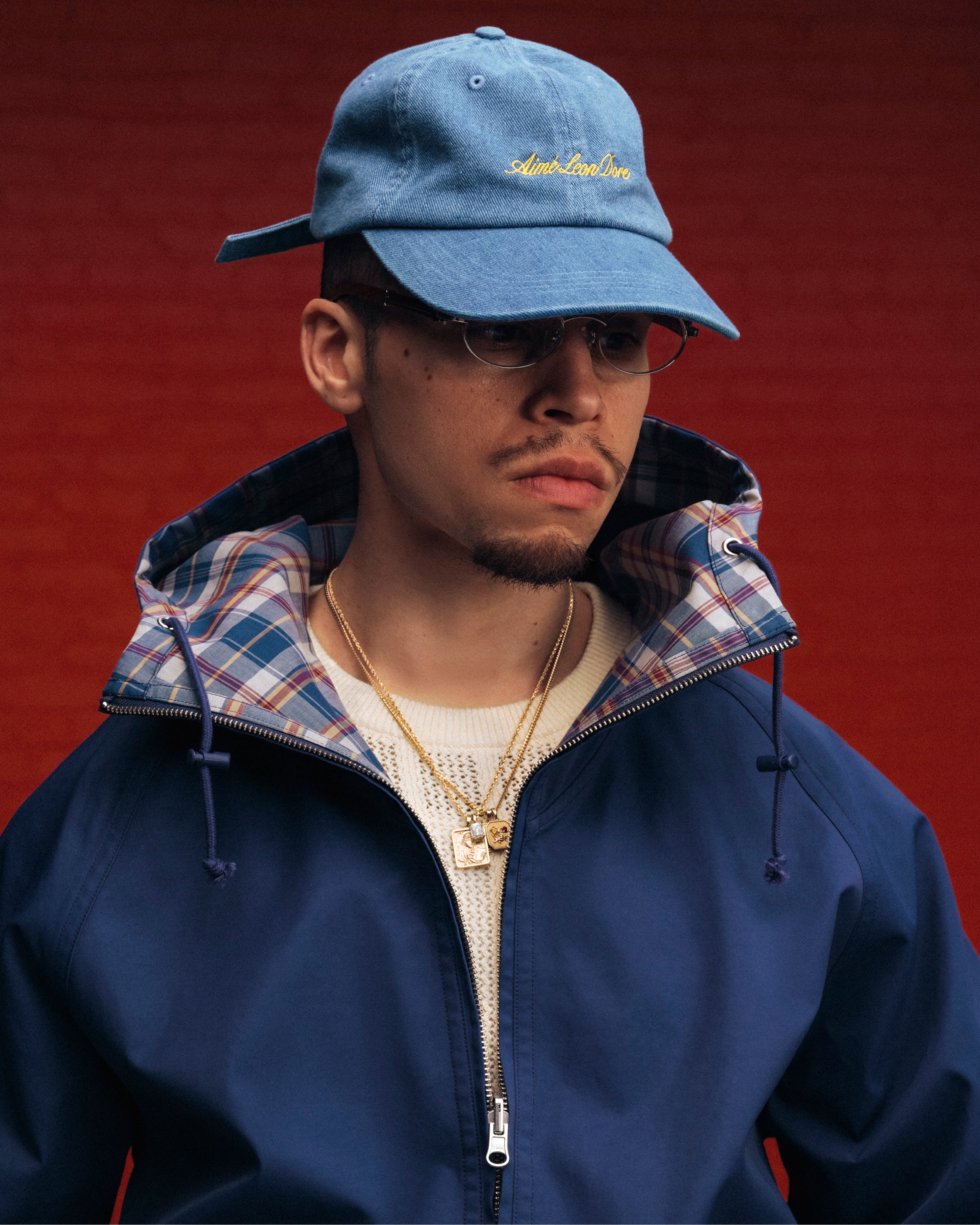 A man in a blue jacket and glasses wears the Denim Script Hat and gold chains, standing against a red background.