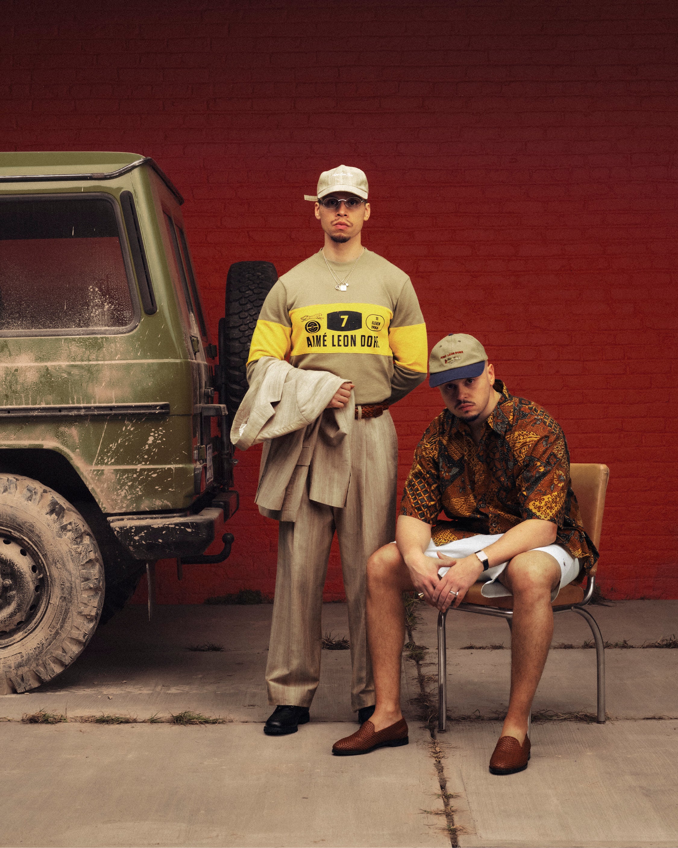 Two men in Double Pleated Safari Suit Trousers pose by a green SUV; one stands, the other sits casually on a chair.