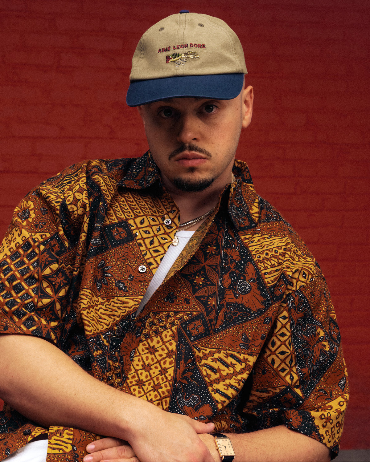 Man in a Printed Abstract Shirt and beige cap sits by a red brick wall, facing the camera.