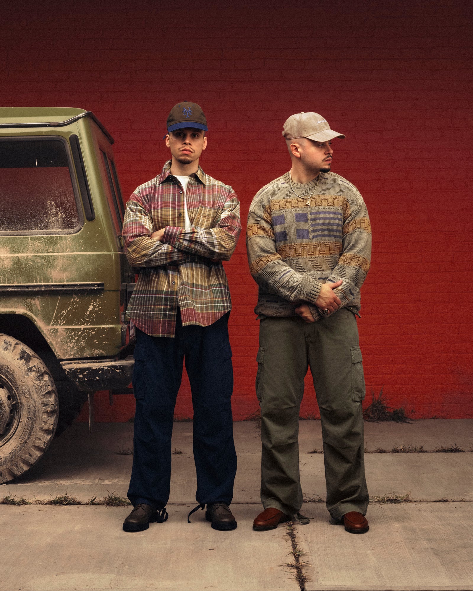 Two men wear Simaía Crewneck Sweaters, standing by a muddy green SUV and a red brick wall, facing forward and sideways.