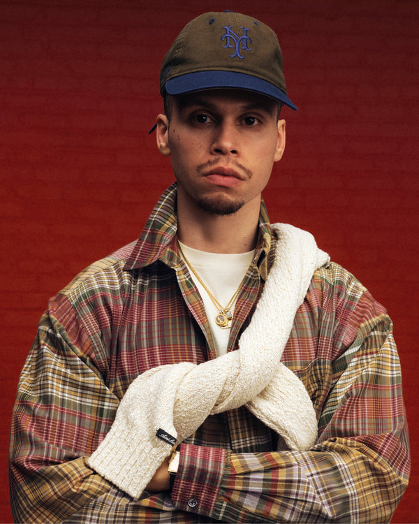 Man in an ALD / New York Mets Colorblock Ballpark Hat and plaid shirt, sweater over shoulders, against a red background.