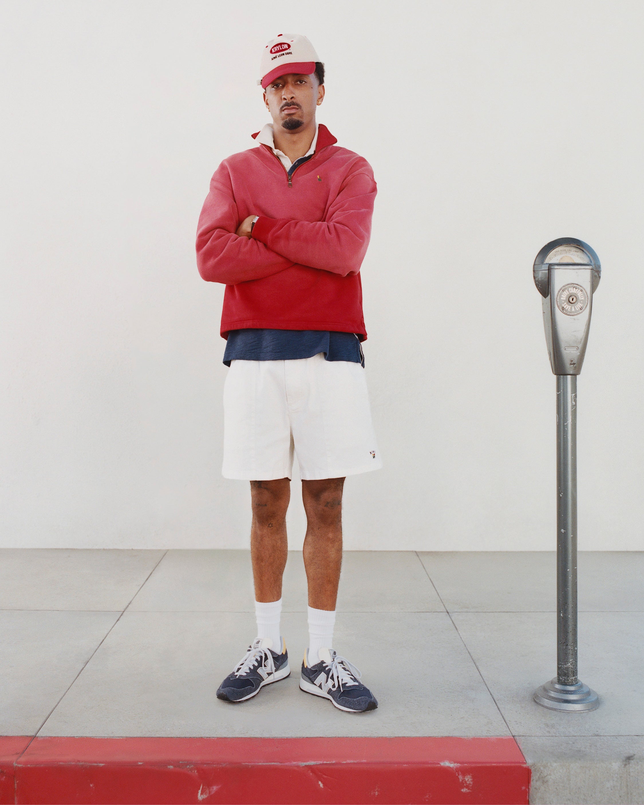 A man in a red jacket and Krylon Logo Hat stands with arms crossed next to a parking meter on the sidewalk.