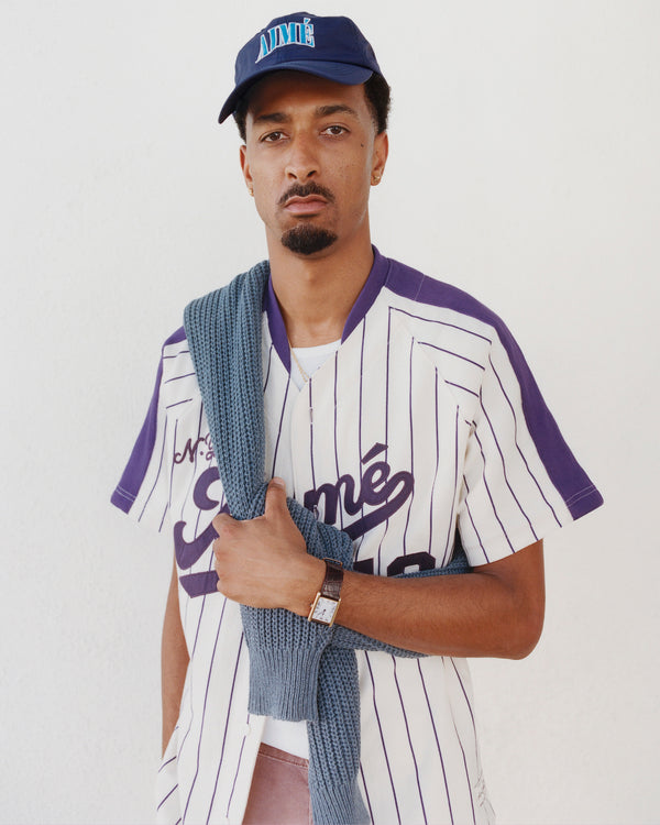 A man in a striped baseball jersey stands against a white wall, wearing a Nylon Crusher Hat and blue sweater over his shoulder.