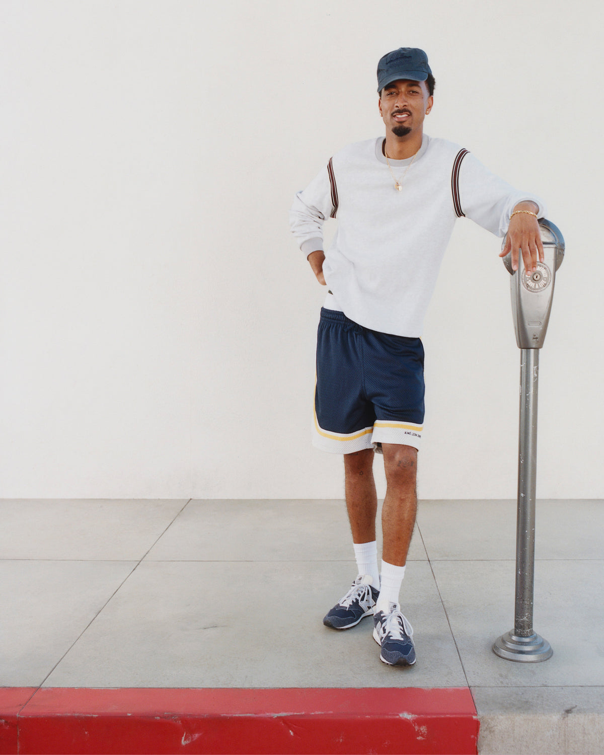 Man in Sun Faded Unisphere Hat, sweatshirt, and shorts leans on parking meter by a white wall.
