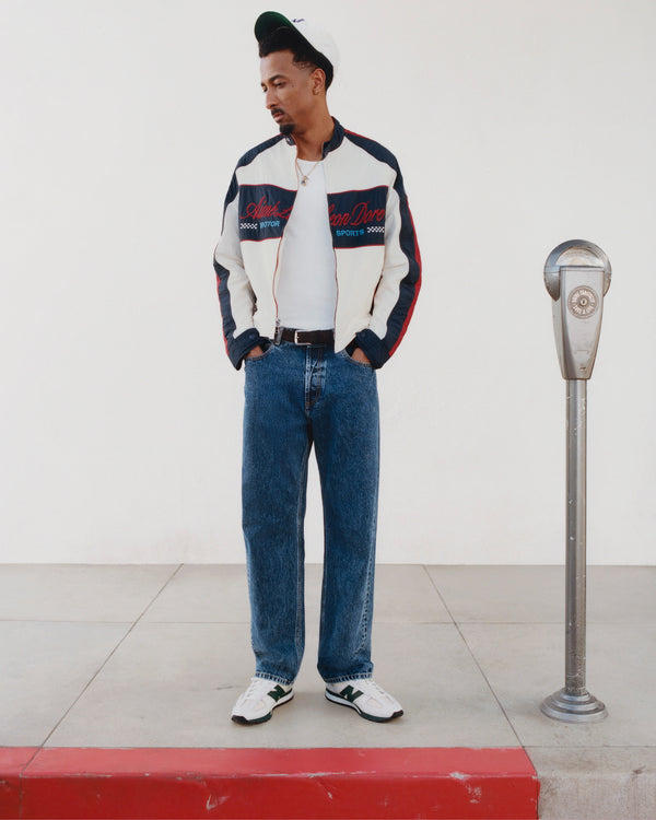 Man in jeans, white shirt, and Nylon Moto Jacket stands on a sidewalk by a parking meter, hands in pockets.