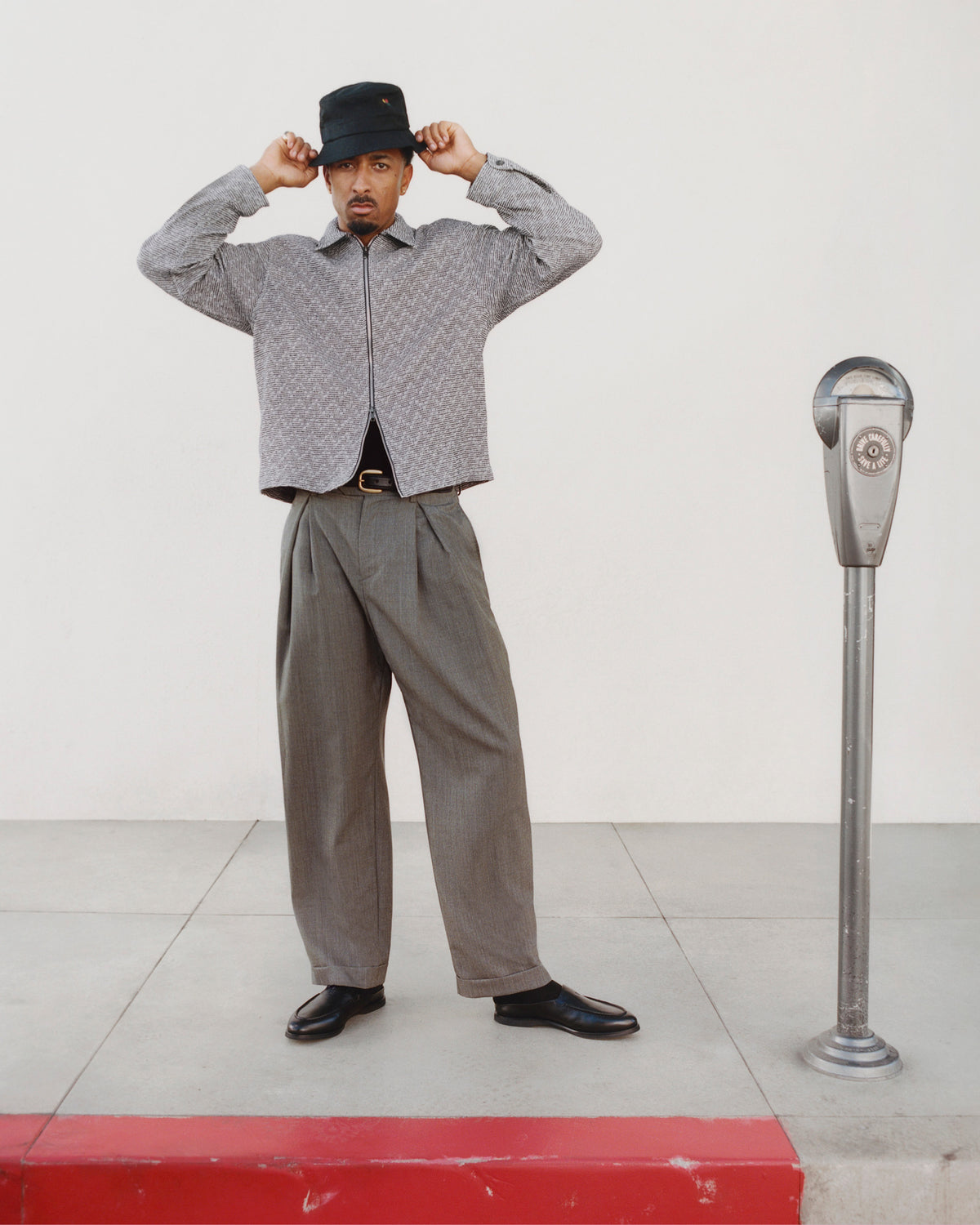 Man in loose gray pants and patterned jacket wears Venetian Mule shoes by a parking meter on the sidewalk.