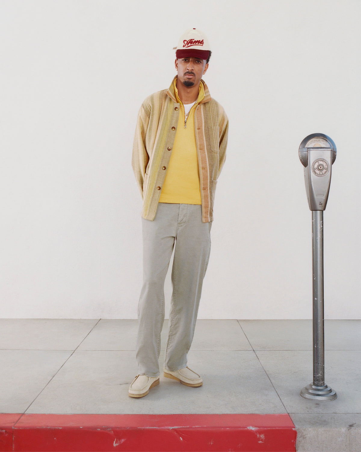 A man in a Blanket Striped Chore Jacket stands on a sidewalk by a parking meter, with a white wall and red curb behind him.