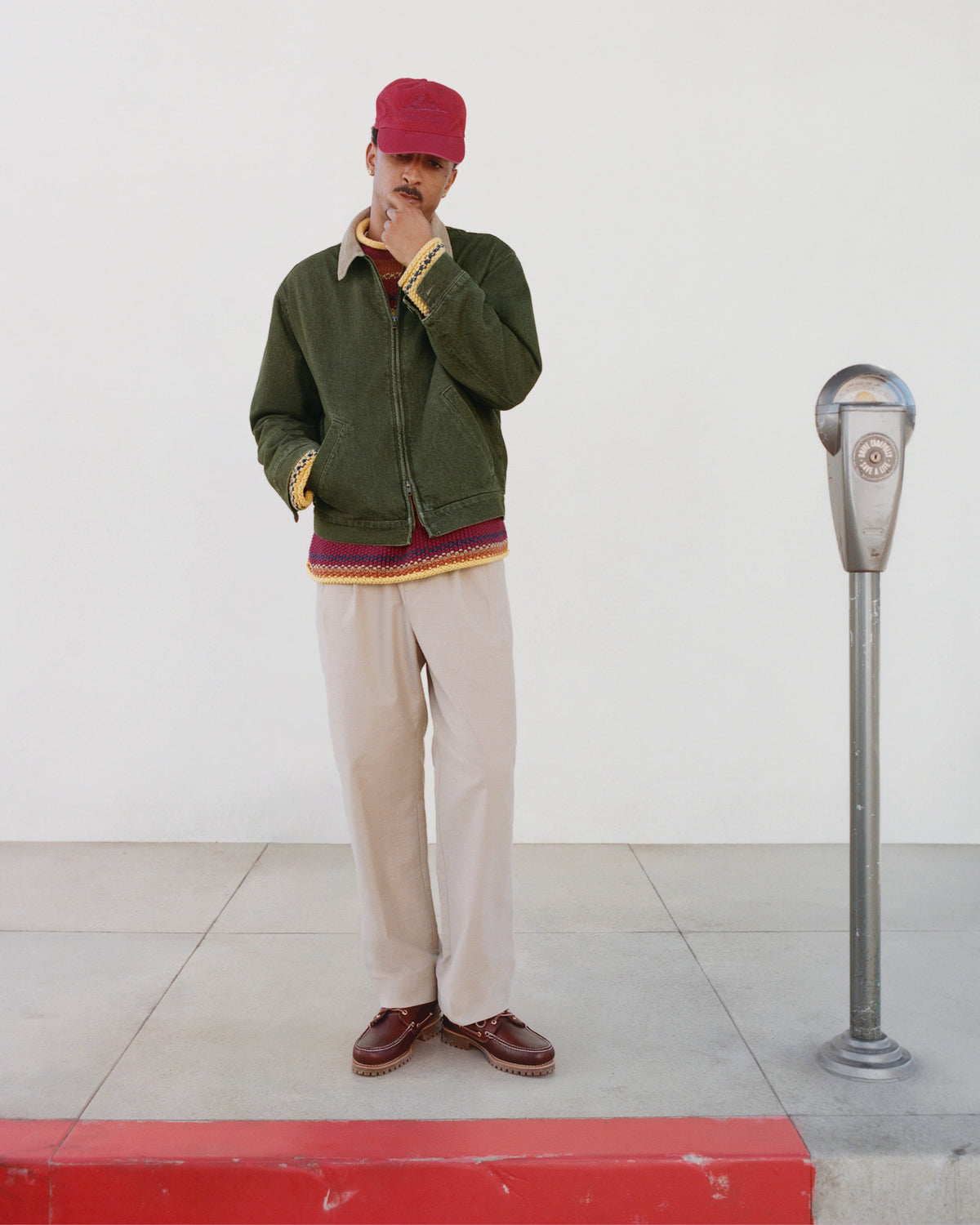 Man in a red cap and Denim Zip Jacket stands by a parking meter on a sidewalk with red curb and white wall.