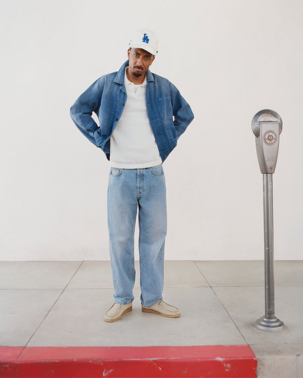 Man in Sun Faded Chore Jacket, white shirt, blue jeans, and LA cap stands by a parking meter on a red-curb sidewalk.
