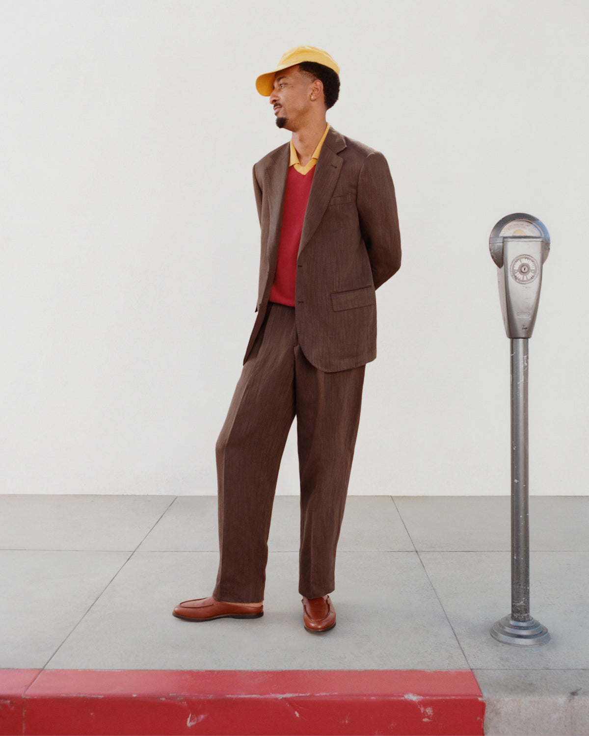 A man in a Single-Breasted Linen Blend Suit Jacket and yellow cap stands by a parking meter on the sidewalk.